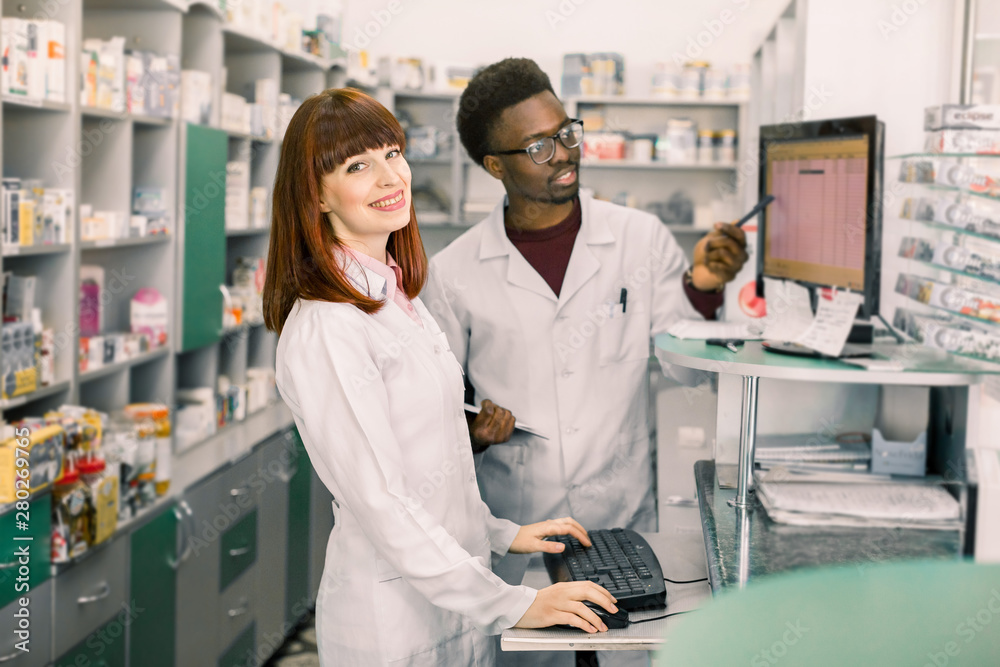 Obraz premium Portrait of a smiling female pharmacist standing in interior of the modern pharmacy, male colleague working with computer in the background.