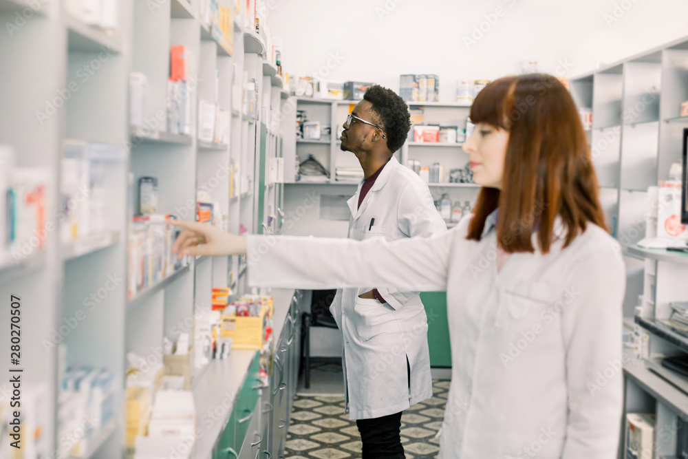 Portrait of a young Caucasian female pharmacist, male African colleague working with drugs in the background. Focus on man