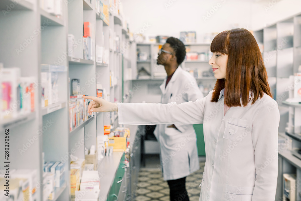 Obraz premium Portrait of a pretty woman pharmacist searching medicines on the shelf at the drugstore, African male colleague working on the background.