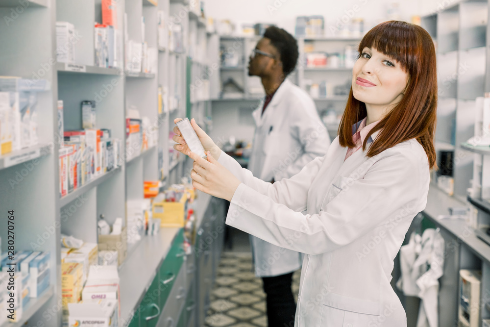Two Pharmacists Working. Caucasian Woman and African Man Wearing Special Medical Uniform. Woman Showing package with Pills. Man looking for medicines on Background.