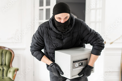 Thief looking at the camera with black balaclava stealing modern Electronic safe box. The burglar commits a crime in Luxury apartment with stucco.