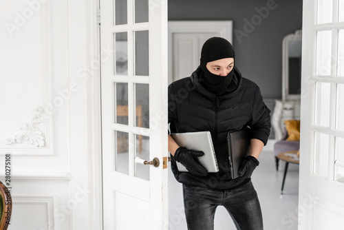 Thief with black balaclava stealing two modern expensive laptops. The burglar commits a crime in Luxury apartment with stucco.