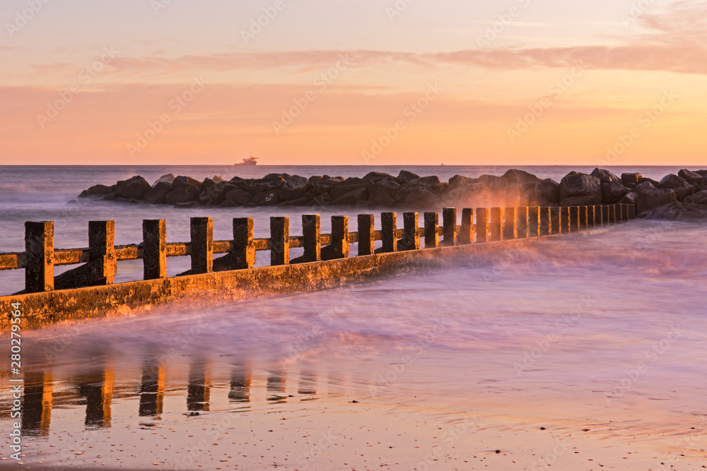 Obraz premium Sunrise at Aberdeen Beach in Scotland. Waves crashing into wooden panels in the water. Beautiful pink sky. Orange, pink color in the sky. Amazing seascape.