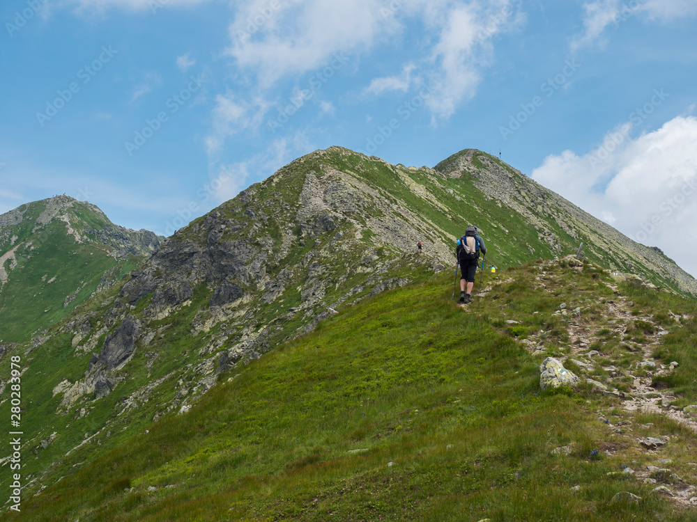 Beautiful mountain landscape of Western Tatra mountains or Rohace with men hiker with backpack hiking trail on ridge. Sharp green grassy rocky mountain peaks. Summer blue sky white clouds.