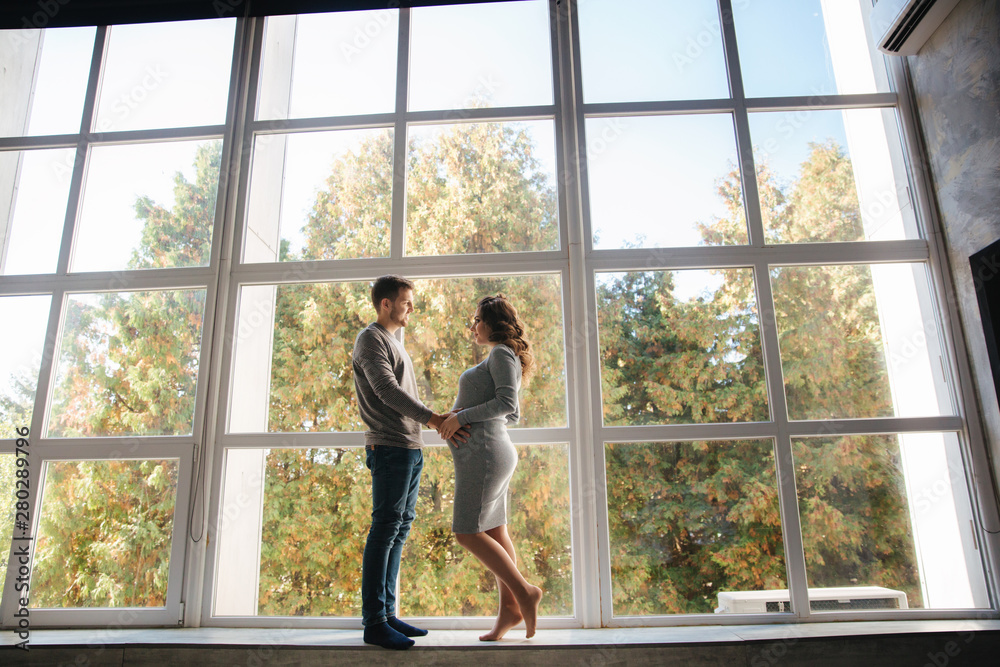 Happy family man and pregnant woman stand in front of big window in their house. They wait for a baby Happy family man and pregnant woman stand in front of big window in their house. They wait for a baby