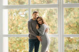 Man hug his pregnant wife and put his hand on her belly. They stand near the window