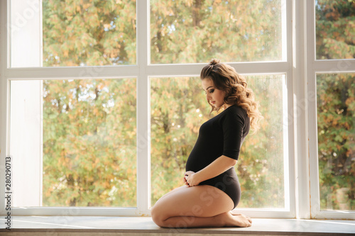 Free Download Pregnant moman sits in front of big window. Model dressed in black bodysuit. She put her hand on the belly. Happy future mother - GFXLoad