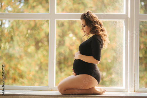 Pregnant moman sits in front of big window. Model dressed in black bodysuit. She put her hand on the belly. Happy future mother
