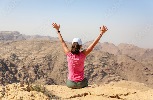 Woman raising her arms overhead in excitement while sitting on a ledge overlooking the desert in Petra. 