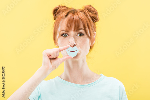 front view of redhead girl holding paper lips and looking at camera isolated on yellow