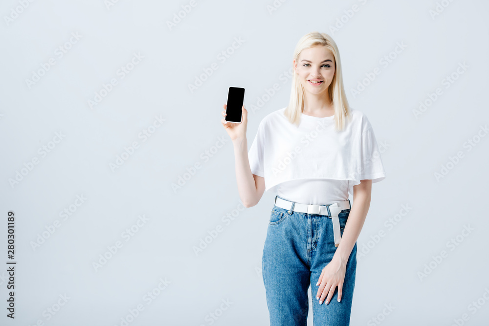 Naklejka premium beautiful happy blonde girl showing smartphone with blank screen isolated on grey