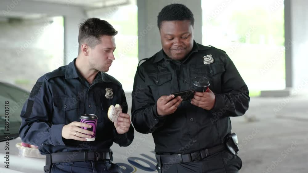 Slow motion two policemen drinking coffee and eating donut use phone talk near patrol car help happy african american break rest smile gangster uniform funny teamwork portrait close up Slow motion two policemen drinking coffee and eating donut use phone talk near patrol car help happy african american break rest smile gangster uniform funny teamwork portrait close up
