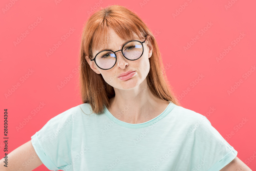 front view of curious redhead woman in glasses looking at camera isolated on pink