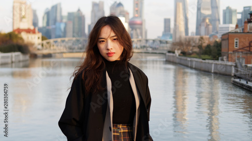 Beautiful young brunette woman in black clothes smiling at camera with  blur Shanghai Bund landmark buildings background in autumn dusk light. Emotions, people, beauty, travel and lifestyle concept.