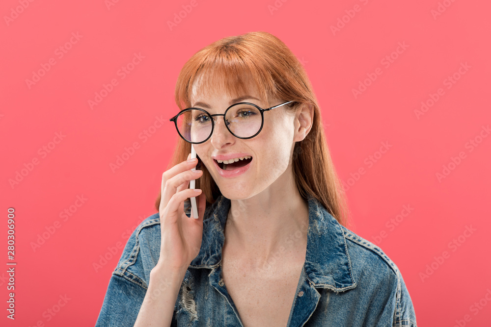 attractive redhead girl in glasses and denim jacket talking on smartphone isolated on pink
