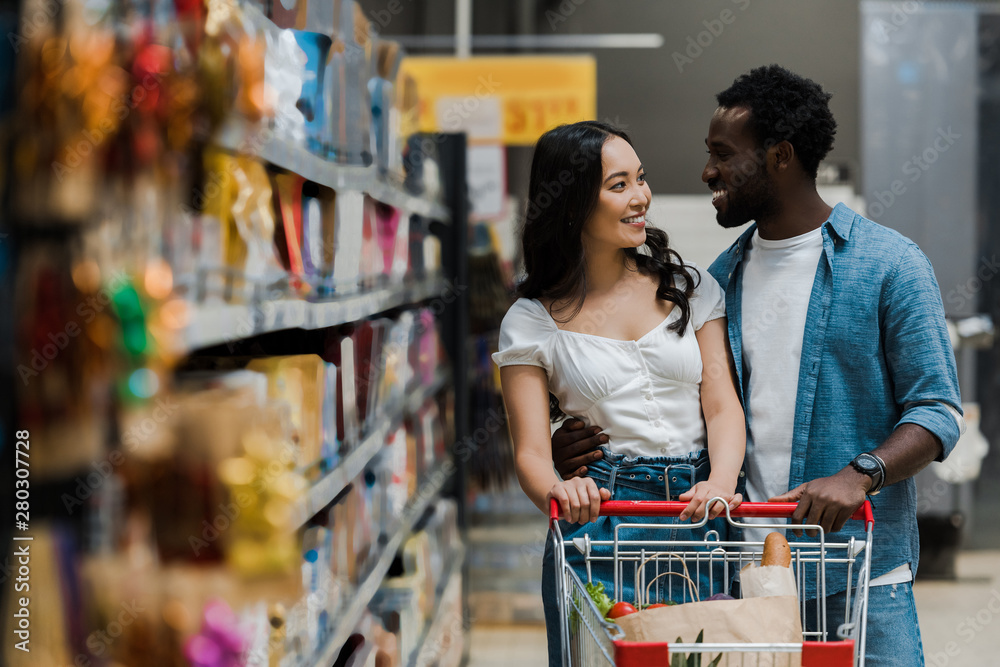 selective focus of happy asian woman looking at cheerful african american boyfriend in supermarket