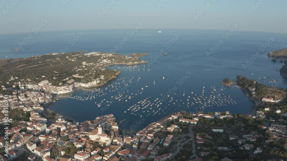 Aerial shot of coastal town Cadaques with bay against scenic seascape. Drone view of Cap de Creus peninsula's coastline, yachts on blue bay, medieval town with tiled roofs and white houses Aerial shot of coastal town Cadaques with bay against scenic seascape. Drone view of Cap de Creus peninsula's coastline, yachts on blue bay, medieval town with tiled roofs and white houses