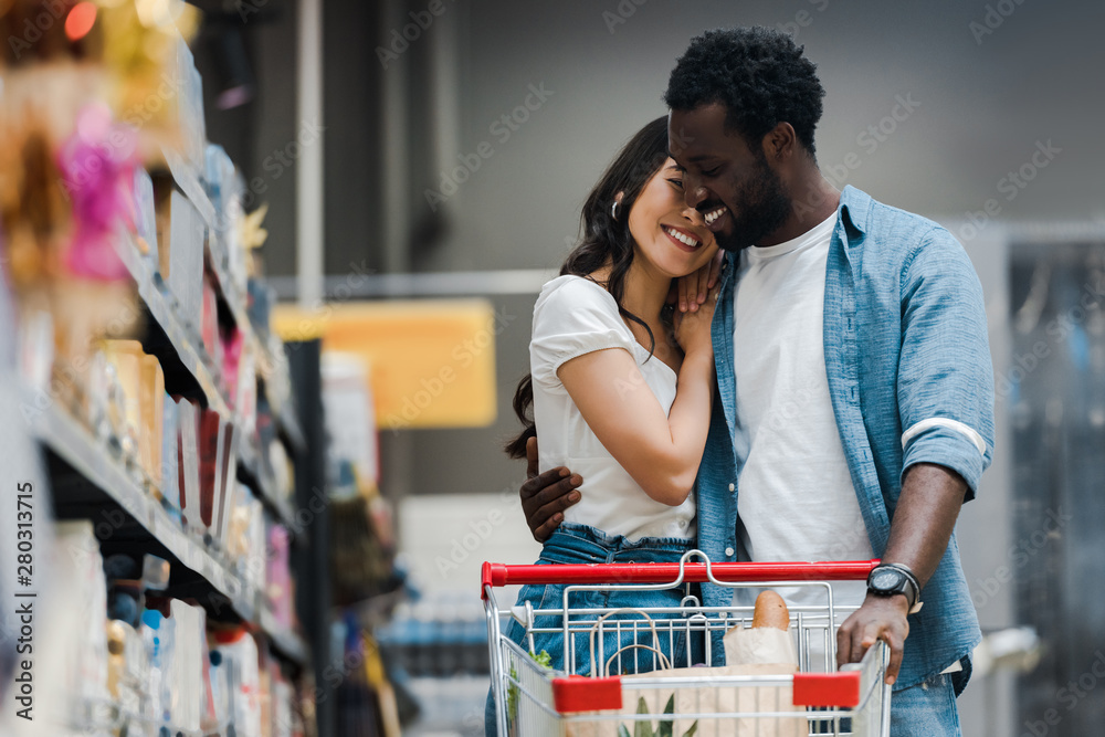selective focus of happy african american man hugging asian girl in supermarket