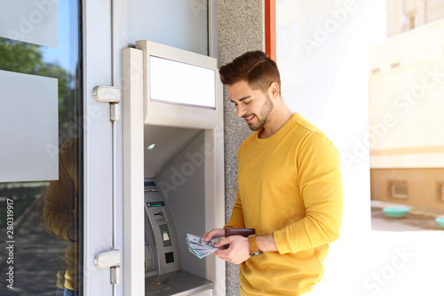 Young man with money near cash machine outdoors