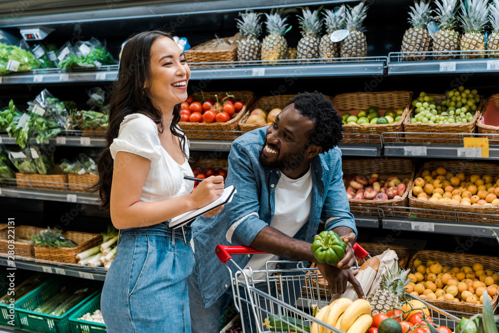 happy african american man looking at asian woman with notebook near shopping cart with groceries