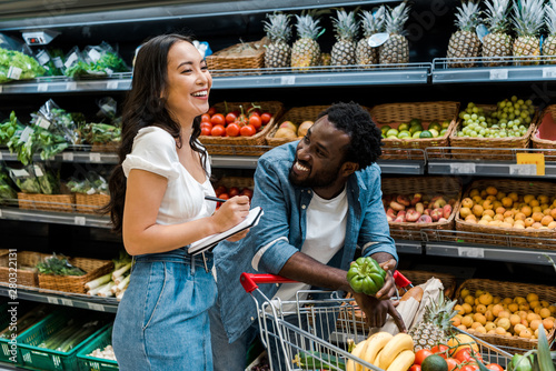 happy african american man looking at asian woman with notebook near shopping cart with groceries photo