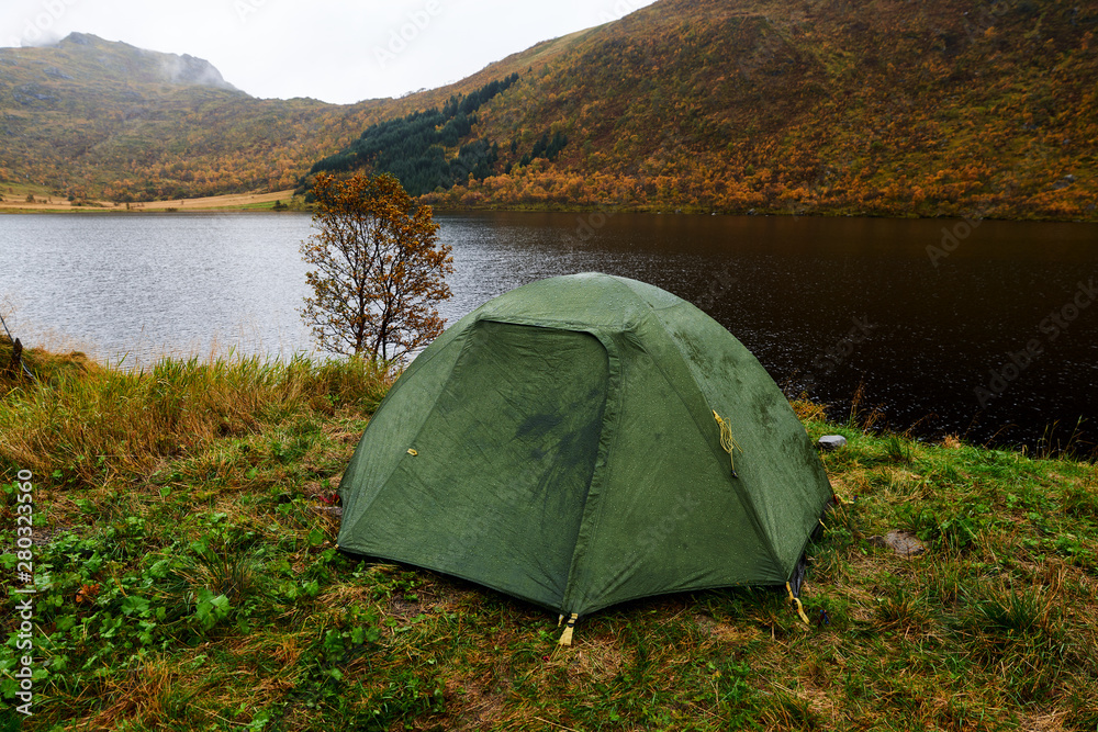 Green tent at a lake surrounded by trees and woodland on Lofoten Islands in Norway