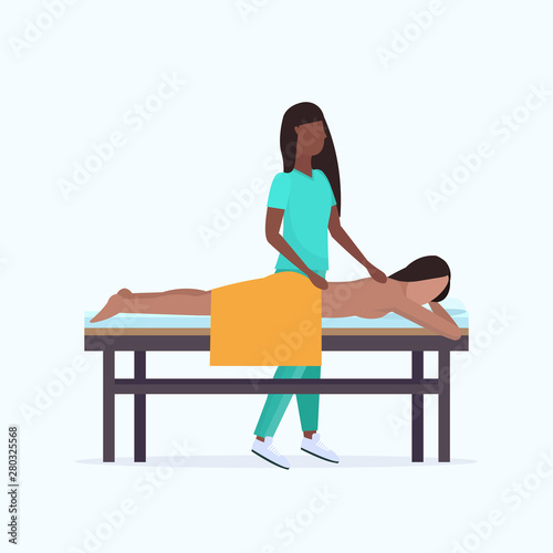 young girl having back massage african american masseuse therapist in uniform massaging patient body woman relaxing lying on bed spa salon treatments concept full length