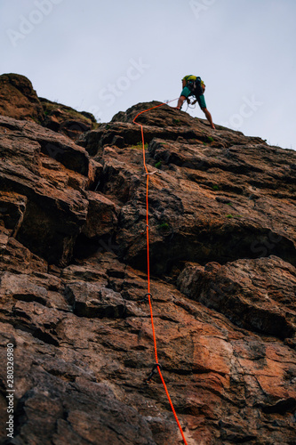 Orange rope and silhouette of a man climbing on cliff against brown cracked rock and blue sky