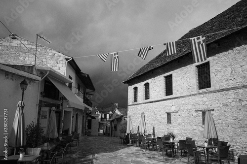Fototapeta Naklejka Na Ścianę i Meble -  Perspective of street with cafe in old town. Architecture in rustic styles. White stone walls and dark dramatic sky. Tables, chairs, umbrellas and flags