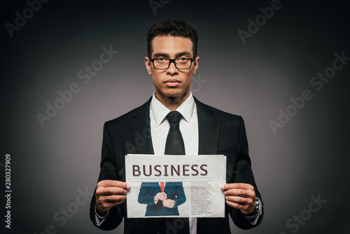handsome african american businessman in glasses and suit holding business newspaper on dark background