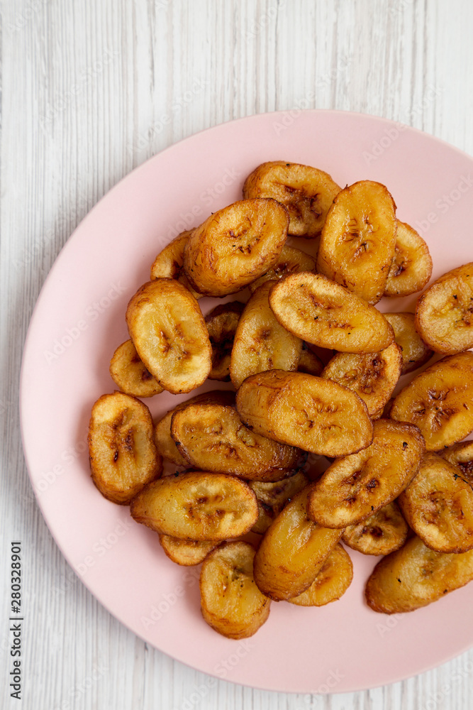 Overhead view, homemade fried plantains on a pink plate on a white wooden surface. Flat lay, overhead, from above. Close-up. Overhead view, homemade fried plantains on a pink plate on a white wooden surface. Flat lay, overhead, from above. Close-up.