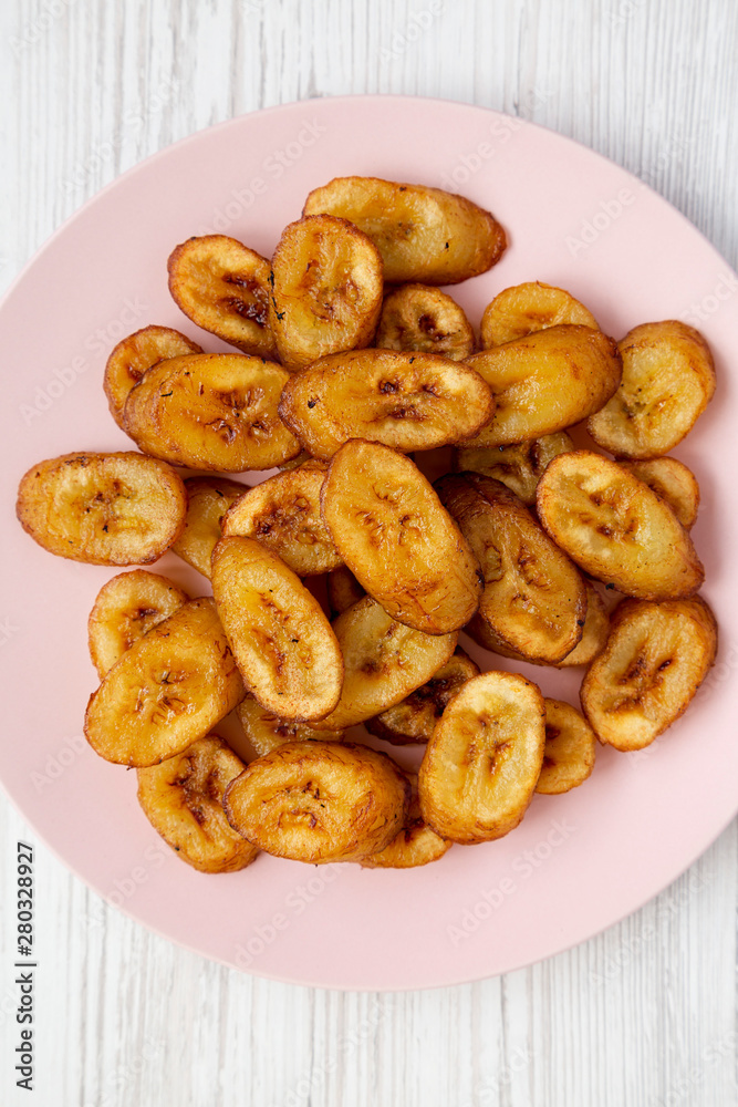Overhead view, homemade fried plantains on a pink plate over white wooden background. Flat lay, overhead, from above. Close-up. Overhead view, homemade fried plantains on a pink plate over white wooden background. Flat lay, overhead, from above. Close-up.