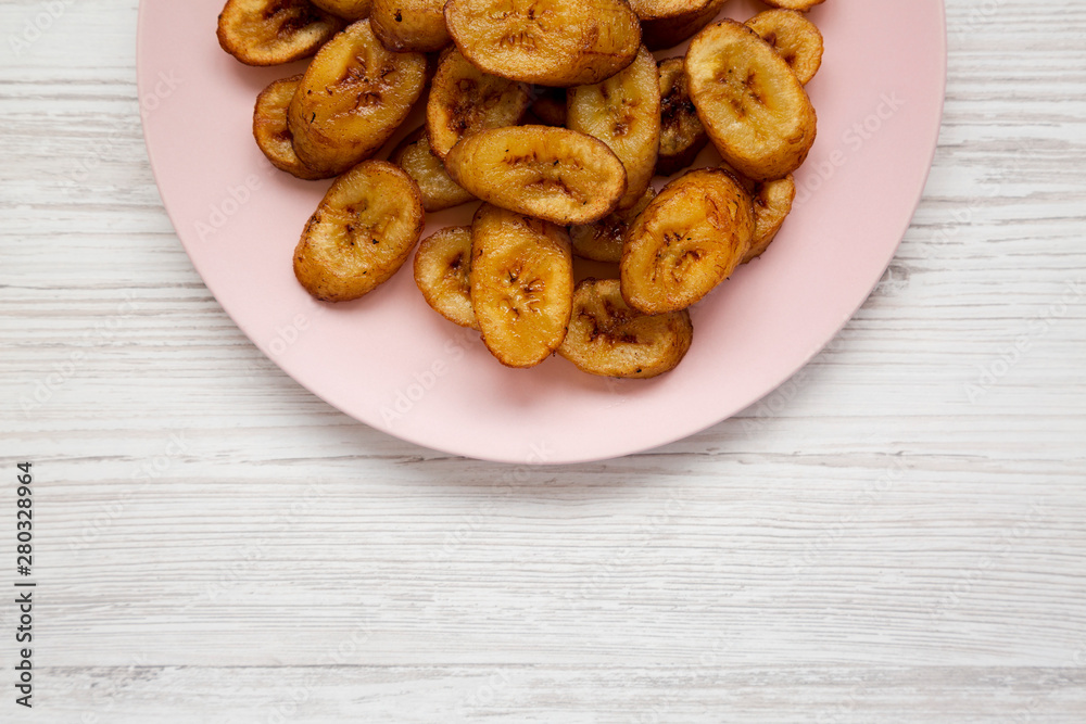 Homemade fried plantains on a pink plate on a white wooden background. Flat lay, overhead, from above. Space for text. Homemade fried plantains on a pink plate on a white wooden background. Flat lay, overhead, from above. Space for text.