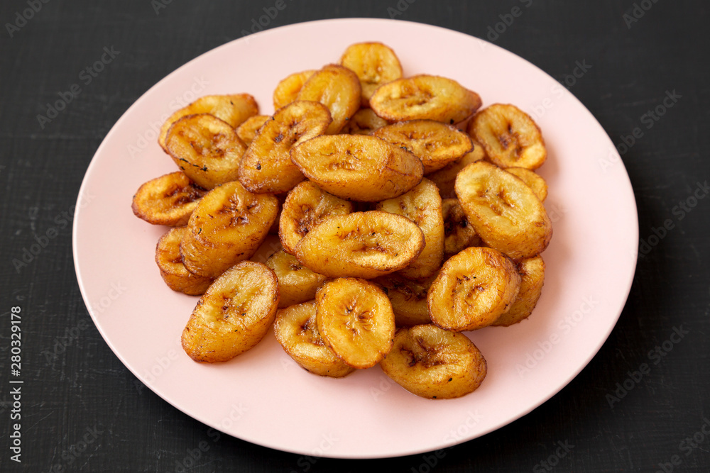 Homemade fried plantains on a pink plate on a black surface, low angle view. Close-up. Homemade fried plantains on a pink plate on a black surface, low angle view. Close-up.