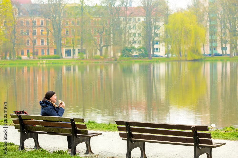 Lady having a sandwich sitting on a bench in a public park in front of a beautiful lake. She is wearing a scarf and a jacket because of the cold weather. Lady having a sandwich sitting on a bench in a public park in front of a beautiful lake. She is wearing a scarf and a jacket because of the cold weather.