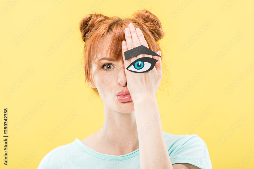 front view of redhead girl holding paper eye and eyebrow and looking at camera isolated on yellow front view of redhead girl holding paper eye and eyebrow and looking at camera isolated on yellow