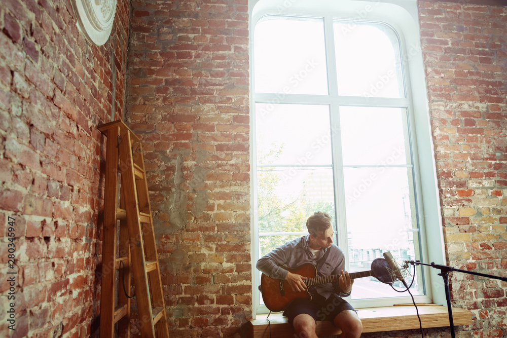 Fototapeta premium Young man recording music video blog, home lesson or song, playing guitar or making broadcast internet tutorial while sitting in loft workplace or at home. Concept of hobby, music, art and creation.
