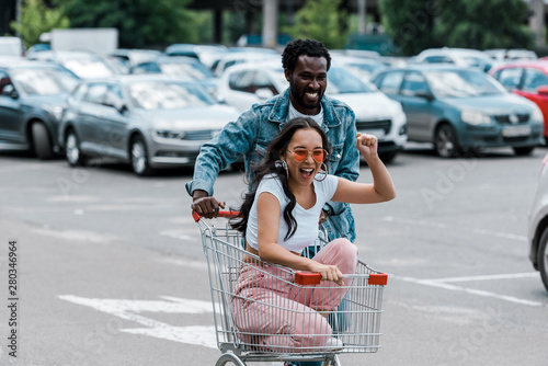 happy asian girl gesturing while sitting in shopping trolley near african american man and cars