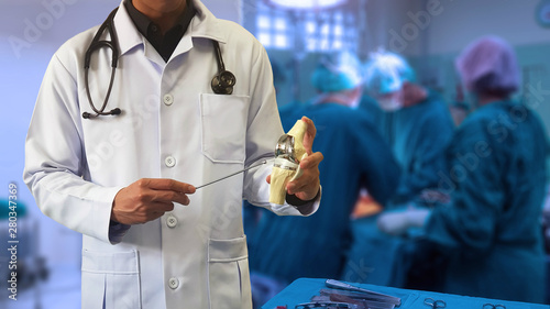 Orthopedic doctor showing model of Total knee replacement with blurred background of surgical team performing surgery in operating room. Medical technology concept. 