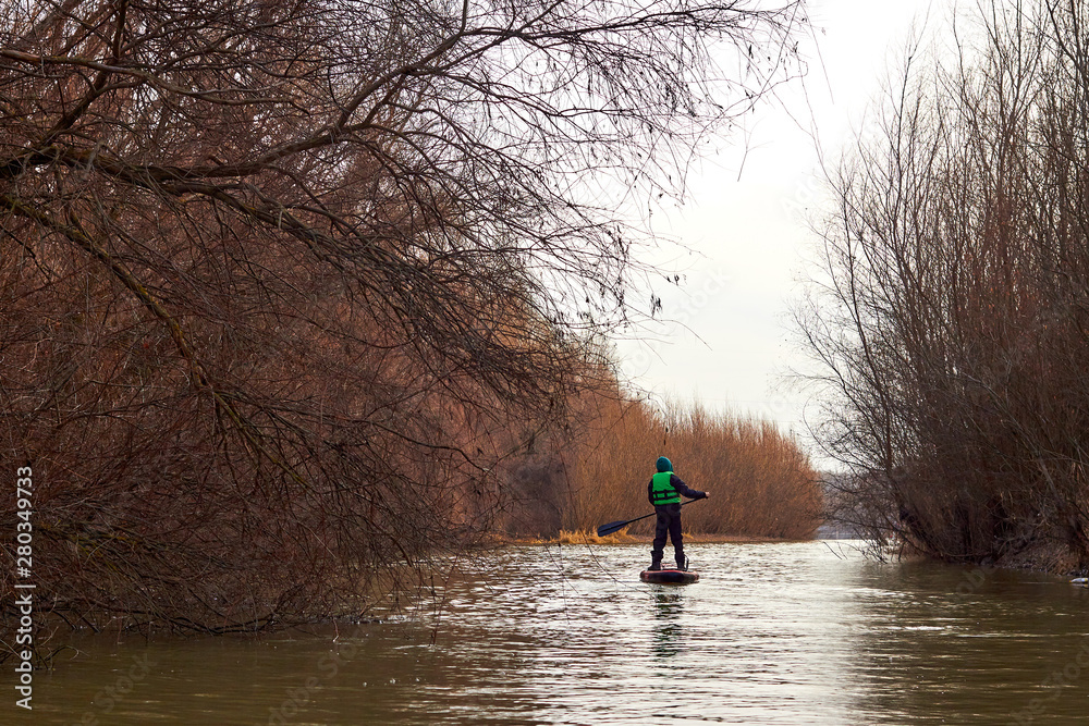 Boy rowing on stand up paddle boarding (SUP) paddling along the calm autumn Danube river against a background of trees at the shore. The concept of children's sports and tourism. Boy rowing on stand up paddle boarding (SUP) paddling along the calm autumn Danube river against a background of trees at the shore. The concept of children's sports and tourism.
