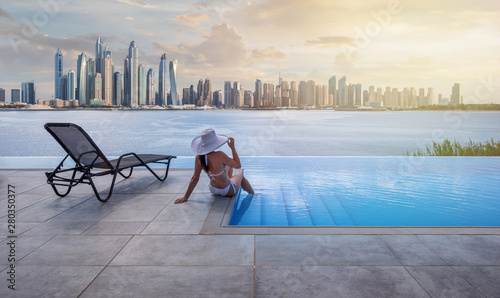 Beautiful panorama of Dubai Marina skyline in a background with a pool, deck chair and woman with a white hat at sunset.