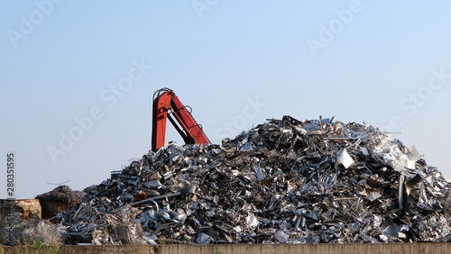 Large stack of aluminum and ferrous materials scrap ready for recycling	shines under the sun. Excavator boom on the left photo