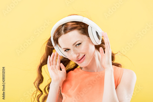 attractive redhead woman listening music in headphones and smiling isolated on yellow