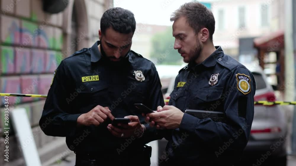 Police at raid. Close-up two handsome policemen searching on smartphones tracking a criminal online. Friendly colleagues cops at police investigation area in the city street.
