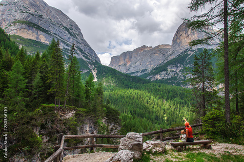 Mother and little daughter sitting on a bench admire a beautiful view of the snow-capped mountains and green spruce in the Dolomites in Italy