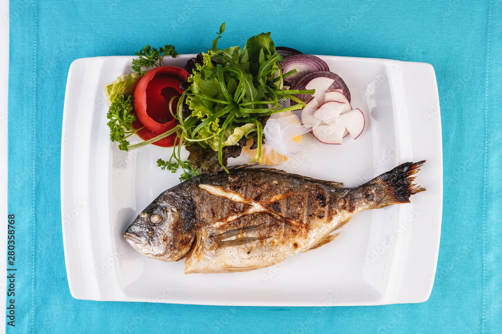 Mediterranean dish, European cuisine. whole fish baked in the oven, served with a salad of vegetables, greens, arugula, onion rings, tomato and lemon Mediterranean dish, European cuisine. whole fish baked in the oven, served with a salad of vegetables, greens, arugula, onion rings, tomato and lemon