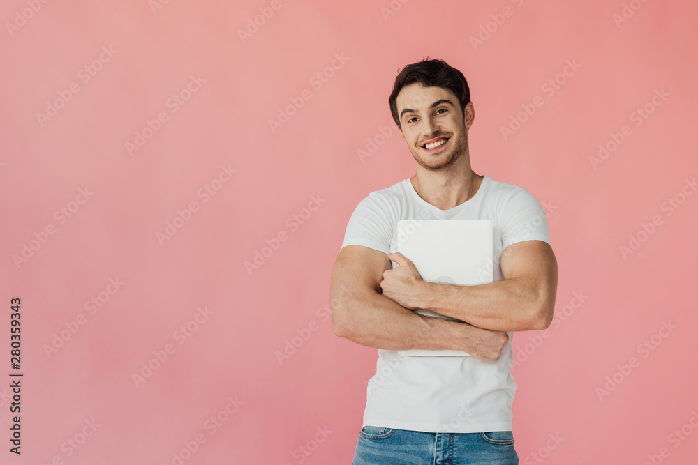 front view of smiling muscular man in white t-shirt holding laptop and looking at camera isolated on pink front view of smiling muscular man in white t-shirt holding laptop and looking at camera isolated on pink