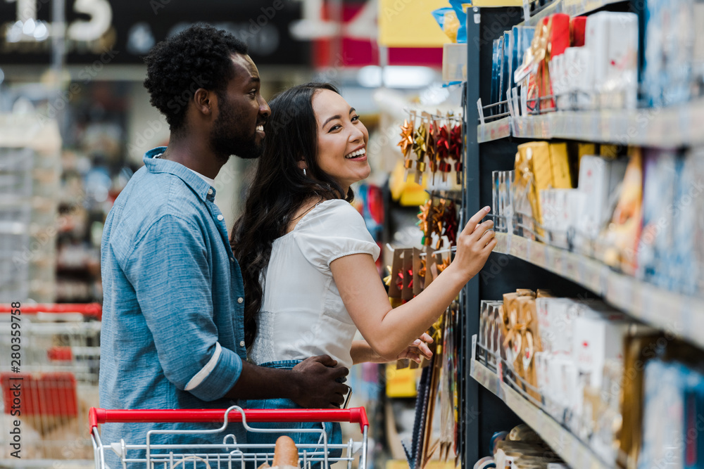 selective focus of cheerful african american man standing with cheerful asian woman pointing with finger at groceries in supermarket