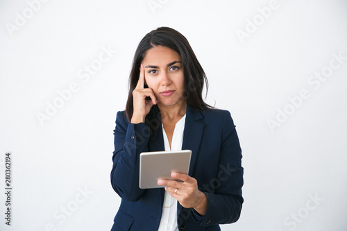 Serious pensive business woman using internet app on tablet. Young Latin woman in office jacket holding digital devise and looking at camera. Online app concept