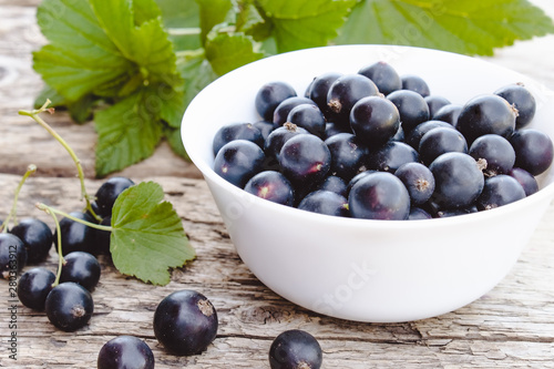 Fresh ripe currant berries in a white bowl on wooden background near green leaves. Juicy fruits currant. Black currant